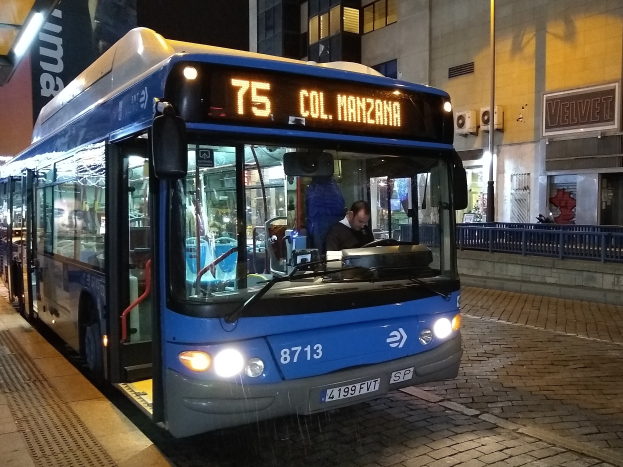 Ein blauer Bus parkt nachts am Straßenrand mit einer Person darin, einer Handtasche im Vordergrund und Gebäuden, Schildern, Geländern, Pfosten und Lichtern im Hintergrund.