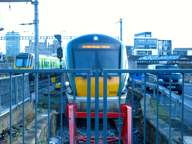 Zwei Züge auf den Schienen mit einem Zaun im Vordergrund, Pfosten auf dem Land, einer mit einer Signalleuchte, ein Auto auf der Straße rechts, Gebäude im Hintergrund und der Himmel darüber.
