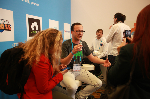 Ein Interview-Szenario mit Menschen, die Mikrofone und eine Kamera halten, ein Banner auf der linken Seite und Personen im Vordergrund und in der Mitte, mit einem klaren Hintergrund.