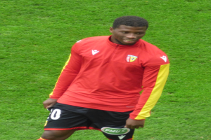 Ein Mann in rotem Trikot und schwarzen Shorts steht auf einem Fußballfeld, lächelt und ist bereit, als Flügelspieler zu spielen.