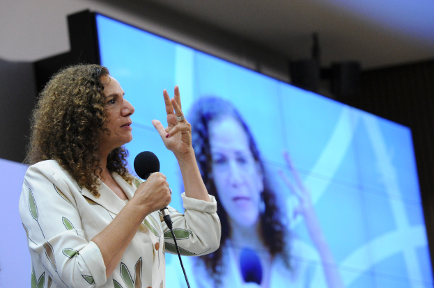 Eine Frau hält ein Mikrofon und spricht, mit einem Bildschirm im Hintergrund, der ihr Bild anzeigt.