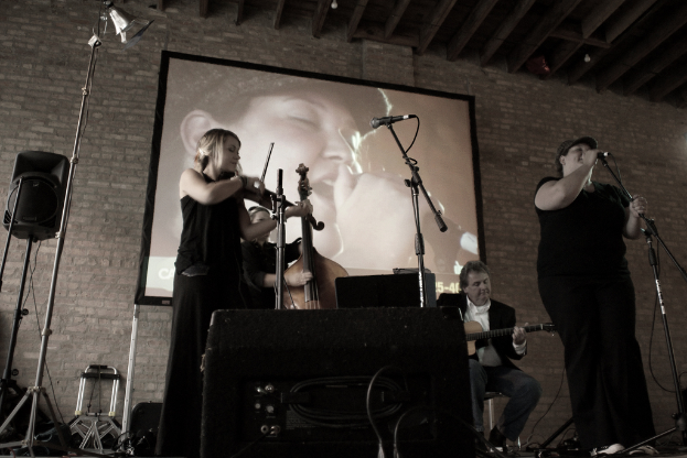 Eine Gruppe von Musikern in Schwarz, mit einem Mädchen, das Geige spielt, zwei Männern, die im Hintergrund Gitarre spielen, und einer Frau, die auf der rechten Seite singt.