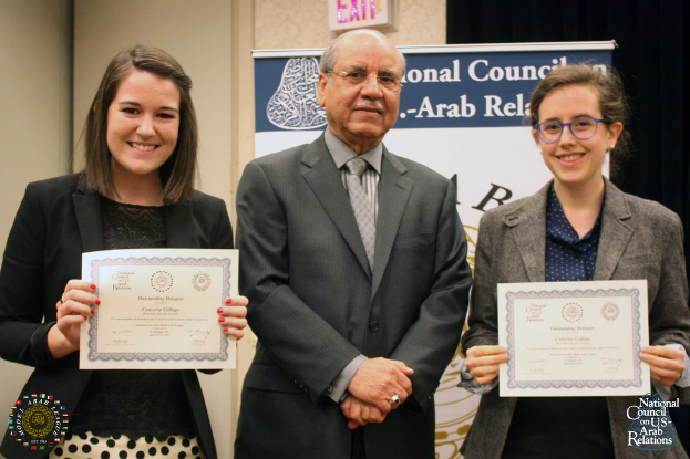 Eine Frau links und eine rechts, beide lachend und Zertifikate haltend, stehen neben einer Person in einem Anzug, mit einer Wandtafel und einem Namensschild im Hintergrund und einem Wasserzeichen im rechten unteren Eck.