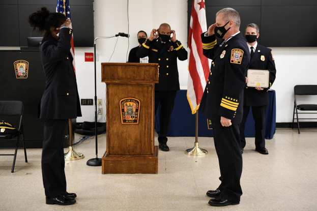 Eine Gruppe uniformierter Personen in Masken steht vor einem Podium mit einem Mikrofon, umgeben von Fahnen, Stühlen und Tischen, während einer Vereidigung eines neuen Feuerwehrchefs, wobei eine Person ein Zertifikat hält.