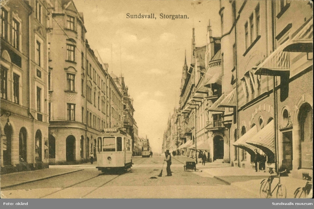 Ein altes Foto einer Stadtstraße mit einer Tram, Gebäuden mit Fenstern, Menschen, die gehen, Fahrrädern und einem bewölkten Himmel, mit Text oben und unten im Bild.