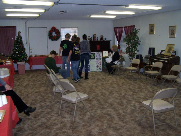 Ein Raum mit mehreren Stühlen, Tischen und Menschen, einige stehen, andere sitzen. Auf den Tischen stehen Becher und Gefäße. Im Hintergrund ist eine Wand mit einem Strauß, einem Vorhang, einer Tür, Rahmen und Fenstern zu sehen. Ein Weihnachtsbaum und ein Piano sind zu erkennen, an der Decke leuchten Röhrenlampen.