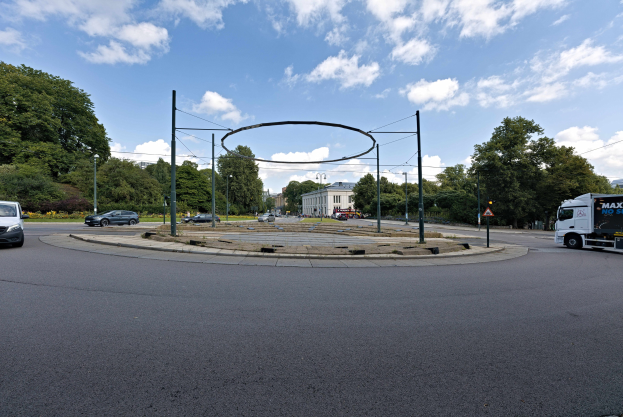 Kreisverkehr mit Fahrzeugen, die darum fahren, umgeben von Pfosten, Bäumen, Gebäuden und einem bewölkten Himmel.