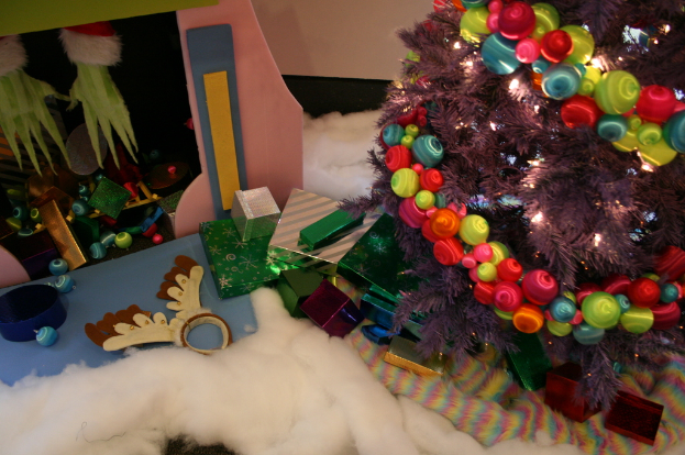 Drinnen steht rechts ein geschmückter Weihnachtsbaum mit Lichterkette und Schmuck, in der Mitte Geschenkschachteln und Dekorationen, oben links eine Hütte mit Dekorationen und unten Wolle und Stoff.