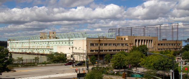 Eine Straße mit Fahrzeugen, Bäumen und Straßenlaternen neben einer Wasserfläche mit einem großen Gebäude im Hintergrund.