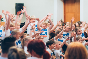 Gruppe von Kindern mit erhobenen Händen vor einer Menge, in der Nähe einer Wand und einer Tür, mit einem Tisch, der eine Vase und einen Lautsprecher hält.
