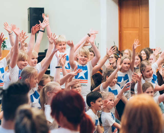 Gruppe von Kindern mit erhobenen Händen vor einer Menge, in der Nähe einer Wand und einer Tür, mit einem Tisch, der eine Vase und einen Lautsprecher hält.