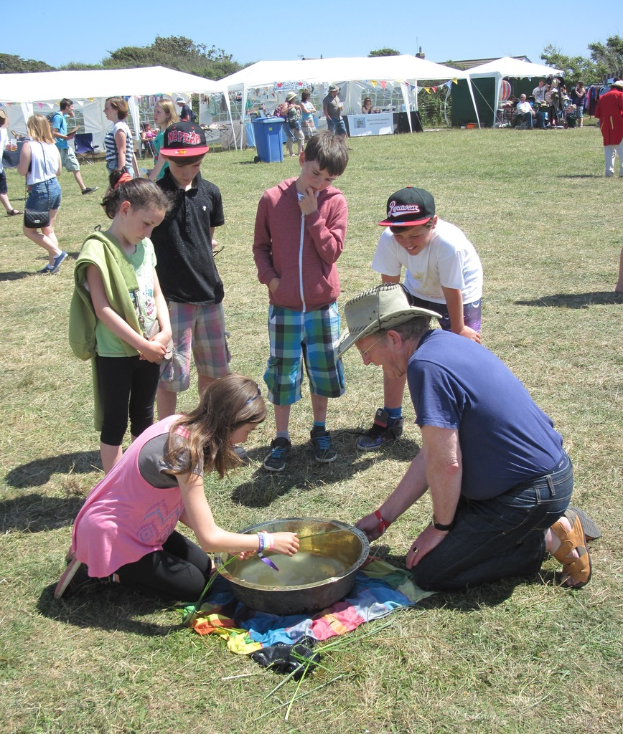 Zwei Personen knien auf dem Boden und arbeiten, mit Kindern im Vordergrund, Zelten, Menschen, einem Müllcontainer und dem Himmel im Hintergrund.