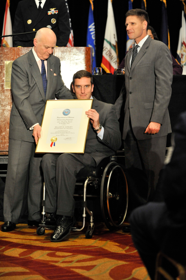 Ein Mann im Rollstuhl hält ein Zertifikat, während zwei Männer in Anzügen auf beiden Seiten von ihm stehen und ihm eine Auszeichnung überreichen, mit einigen Menschen, Flaggen und einem Podium im Hintergrund.