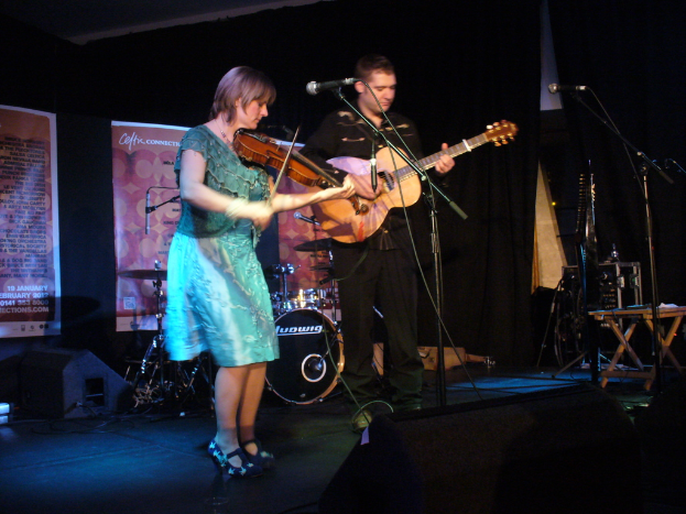 Ein Mann in schwarzem Hemd und Hose spielt Gitarre in der Mitte der Bühne, flankiert von einer Frau in einem blauen Kleid, die Geige spielt, sowie verschiedenen Musikinstrumenten und Lautsprechern im Hintergrund.