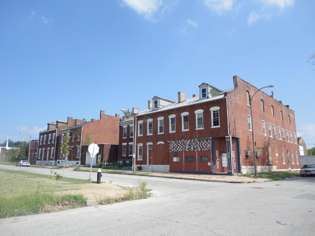 Ein Gebäude mit Masten und Straßenlaternen, umgeben von Bäumen und Fahrzeugen auf beiden Seiten, mit einem Weg, Gras und einem Feuerlöscher unten und dem Himmel oben.