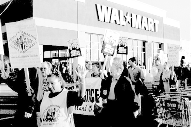 Ein Schwarz-Wei├č-Bild von protestierenden Menschen, die Plakate halten, mit einem Einkaufszentrum im Hintergrund.
