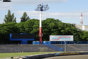 Rennstrecke mit Überdachung, umgeben von Gras, Geländern, Stufen, einem Lichtmast, einem Schild, Bäumen, Gebäuden, Türmen und einem bewölkten Himmel.