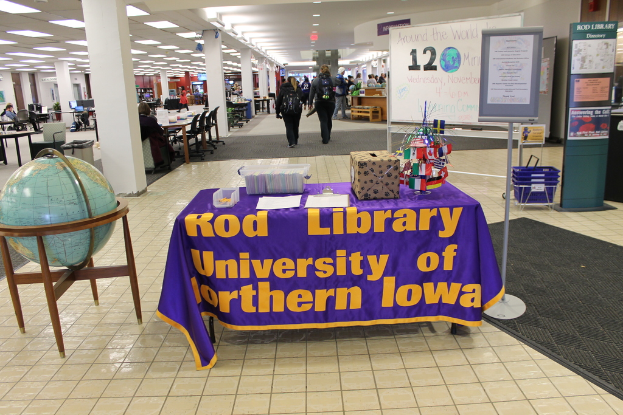 Eine Ausstellungssituation mit einem Tisch, auf dem verschiedene Landesflaggen, eine Box, Papiere und ein Banner liegen, eine Kugel links daneben, ein Anschlagbrett rechts daneben mit ein paar Menschen daneben und mehreren Tischen und Stühlen links.
