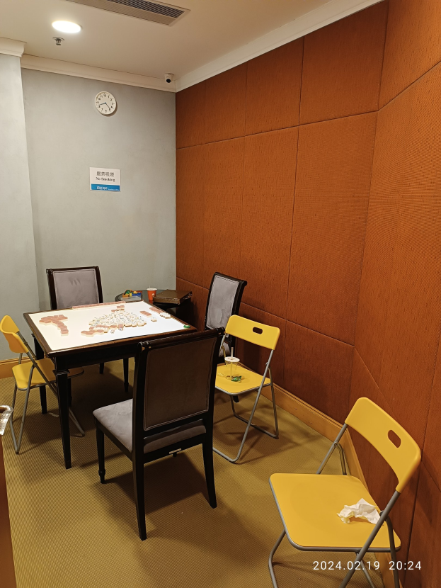 Ein Raum mit einem Tisch, Stühlen, einer Uhr an der Wand, einem Papier an der Tür, Deckenleuchten, verschiedenen Gegenständen auf dem Tisch und einem nahen Müllbehälter.