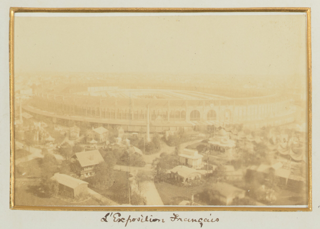 Schwarz-weißes Foto eines Stadions in städtischer Umgebung, umgeben von Gebäuden, Bäumen und Strommasten, mit Text unten.