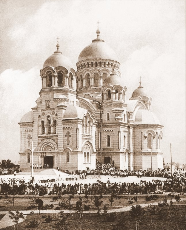 Schwarz-weiß-Fotografie der Erlöserkirche auf dem Bluten Blut in St. Petersburg, Russland, mit Menschen, die sich um das prunkvolle Gebäude versammeln, Bäumen und Pfählen im Vordergrund und einem klaren Himmel im Hintergrund.