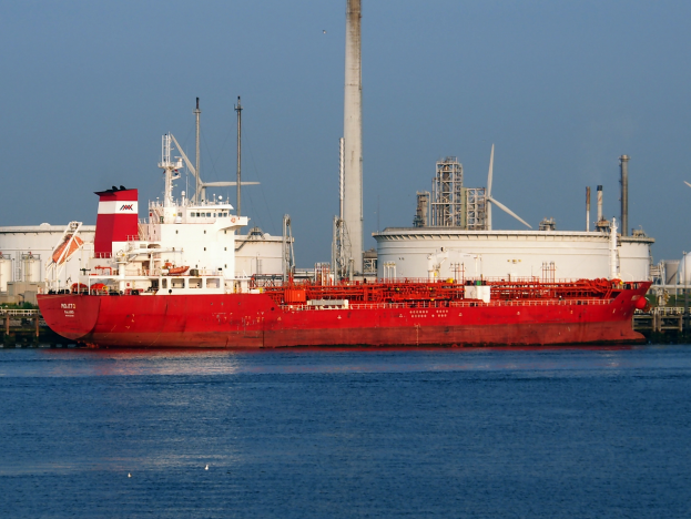 Ein großes rotes und weißes Öltankschiff, das auf einem Gewässer schwimmt, mit einer Fabrik im Hintergrund und einem klaren blauen Himmel.