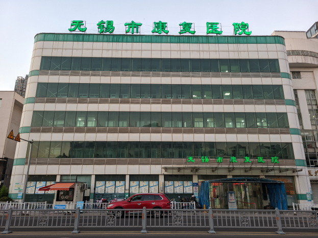 Ein großes Gebäude mit vielen Fenstern, das als chinesisches Krankenhaus bezeichnet wird, ist von einem Metallzaun umgeben, mit Fahrzeugen auf der Straße, Menschen auf dem Bürgersteig, einem Schild, Pflanzen und einem bewölkten Himmel.