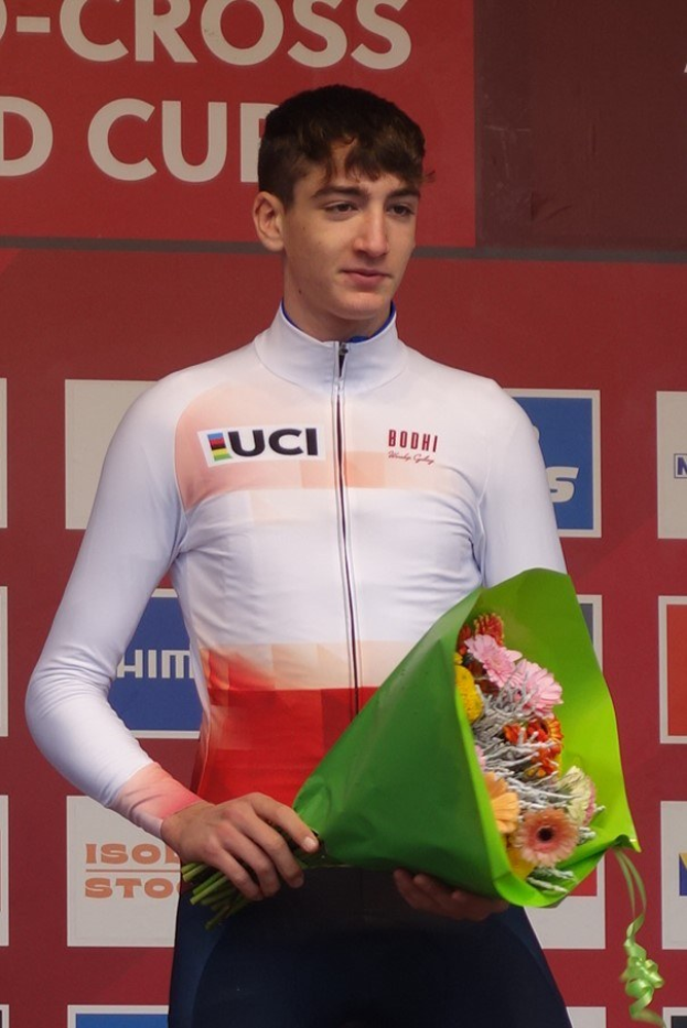 Ein Mann in einem Anzug steht selbstbewusst auf einem Podium und hält einen Blumenstrauß in der Hand, mit einem Siegerboard im Hintergrund.
