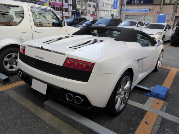 Ein weißer Audi R8 Spyder steht in einer Parkfläche mit anderen Autos, umgeben von Gebäuden, Schildern, Pfählen, Bäumen und einem klaren blauen Himmel.