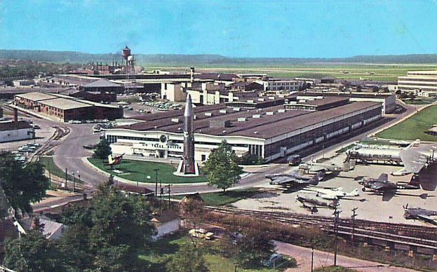 Alte Schwarz-Weiß-Airport-Szene mit geparkten Propellerflugzeugen, Terminalgebäuden, Bäumen, Straßen, Fahrzeugen, Versorgungsmasten, Gras, Bahnschienen, fernen Türmen, rollenden Hügeln und einem klaren Himmel; Bildunterschrift erscheint unten.