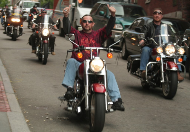 Eine Gruppe von Menschen, die auf Fahrrädern auf einer Straße fahren, wobei eine Person in einem roten Hemd in der Mitte ohne Hände auf dem Lenker fährt, eine Person in einer schwarzen Jacke rechts lächelt und zwei Autos und ein Van im Hintergrund zu sehen sind.