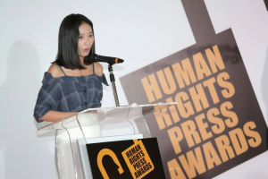 Eine Frau in einem schwarzen Kleid mit einem Dutt steht an einem Podium mit einem Mikrofon und hält eine Rede vor einem Banner 'Human Rights Press Awards', ihr Gesichtsausdruck ist ernst und konzentriert.