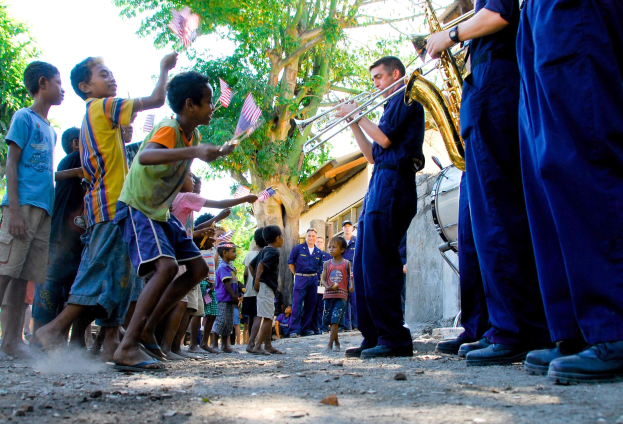 Gruppe von Kindern, die Musikinstrumente im Freien spielen, mit einer Menge, die Fahnen schwingt, im Hintergrund, Bäume, Gebäude und einen klaren blauen Himmel.