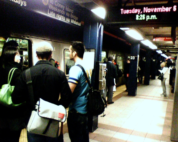Eine volle Bahnsteig mit Menschen mit Taschen, ein Zug auf der linken Seite und ein Textschild oben.