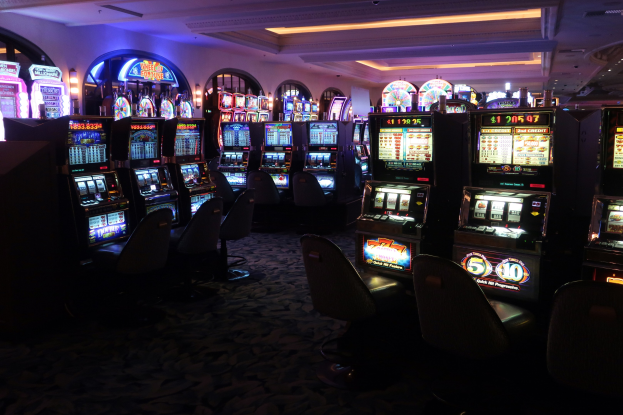 Eine Reihe von Spielautomaten mit Text und Zahlen darauf, die in einem Casino mit Stühlen auf dem Boden und Deckenlampen angeordnet sind.