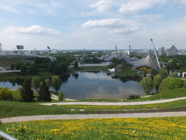 Ansicht des Olympiageländes von einem Hügel aus, mit einem Teich im Vordergrund, umgeben von Grün und gelben Blüten, und im Hintergrund Gebäude und Masten gegen einen klaren blauen Himmel.