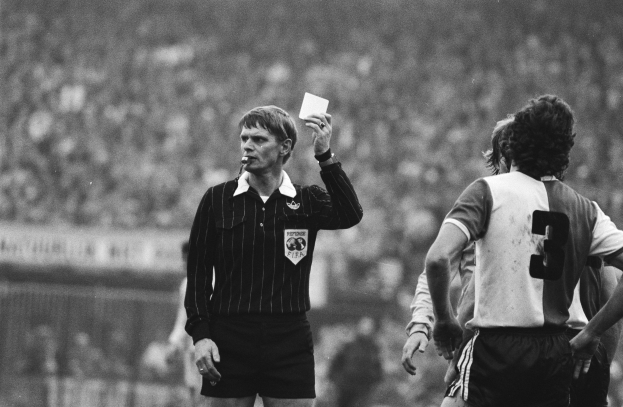 Schwarzes und weißes Bild eines Schiedsrichters mit roter Karte, mit einer Menge Menschen in Fußballuniformen im Hintergrund.