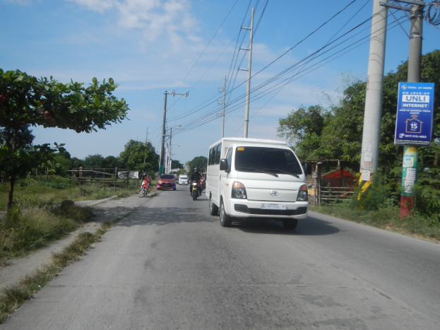 Ein weißer Lieferwagen fährt eine Straße entlang, die von Strommasten und Drähten gesäumt ist, mit Fahrradfahrern in der Nähe, Bäumen und Gras auf beiden Seiten, unter einem klaren blauen Himmel.