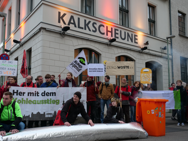 Eine Gruppe von Menschen mit Schildern und Plakaten steht vor einem Gebäude, zwei Personen sitzen im Vordergrund und ein Müllcontainer rechts, während einer Demonstration in Deutschland.