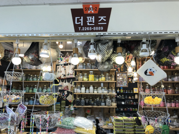 Ein Ladeneinrichtung mit verschiedenen Gegenständen wie Körbchen, Lichtern, Flaschen und Spielzeugen, die auf Gestellen angeordnet sind, mit einer Tafel öber dem Eingang, die es als bestes koreanisches Geschäft in Seoul ausweist.