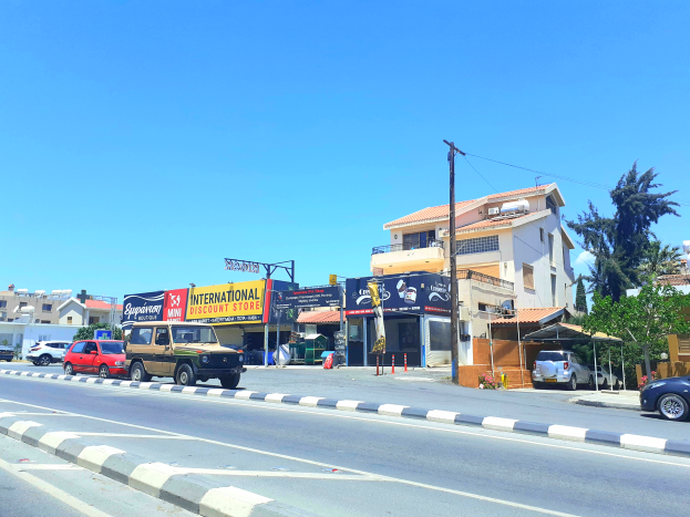 Eine Straßenszene mit parkenden Autos, einer Begrenzung, Verkehrskegeln, Pfählen und Schildern mit der Aufschrift "Istanbul International Discount Store", mit Gebäuden, Bäumen und einem bewölkten Himmel im Hintergrund.