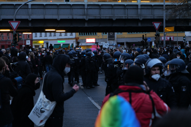 Eine große Gruppe von Menschen steht vor einer Reihe von Polizeibeamten, einige tragen Helme und tragen Taschen oder Kameras, mit Gebäuden, Schildern, Laternen, Verkehrsampeln, einer Brücke und einem Baum im Hintergrund.