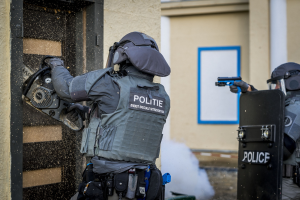 Zwei Polizisten in Einsatzkleidung stehen vor einem Gebäude, einer hält eine Waffe und der andere ein Objekt, mit einer Tür links und einer Tafel an der Wand im Hintergrund.