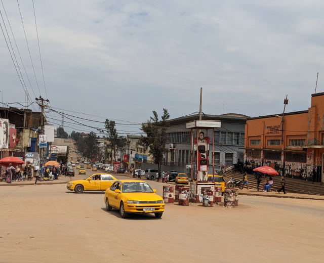 Ein gelbes Taxi fährt eine belebte Stadtstraße mit Gebäuden und Fenstern an den Seiten hinunter, während Fußgänger mit Regenschirmen auf dem Gehweg unter einem bewölkten Himmel gehen, mit Strommasten und Kabeln im Hintergrund.