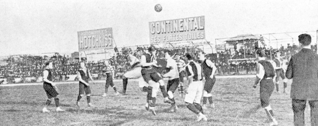 Ein Schwarz-Weiß-Bild einer Gruppe von Männern, die auf einem Feld Fußball spielen, mit einem Ball in der Luft und einer Zuschauermenge im Hintergrund, mit Tafeln, die Text über das Spiel anzeigen.