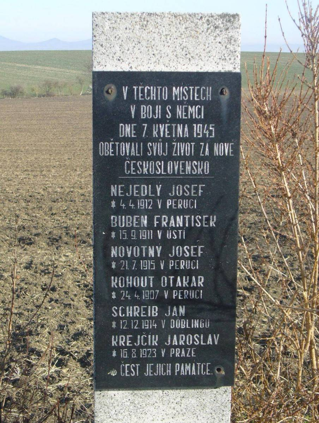 Ein Denkmal mit einer Tafel, auf der "V Techno Mistech" steht, befindet sich inmitten eines Feldes, umgeben von Pflanzen, Hügeln und einem klaren blauen Himmel.