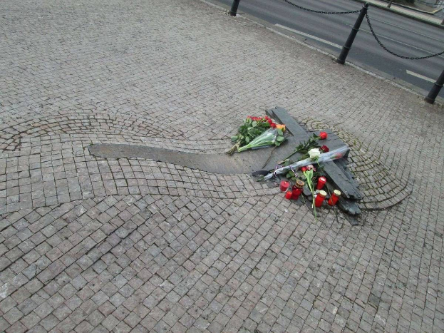Gedenken mit Blumen, Kerzen und verstreuten Buketts am Straßenrand, umgeben von Bäumen, Gebäuden und Fahrzeugen.