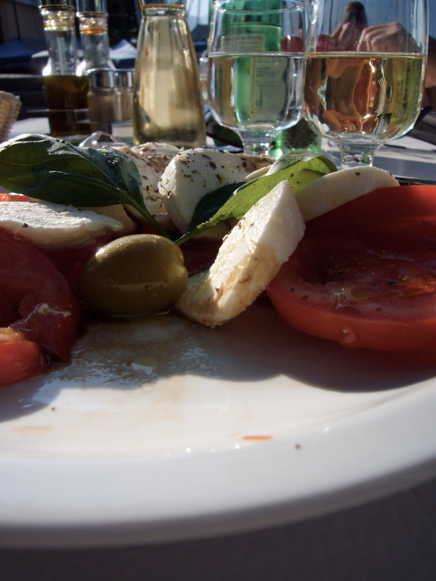 Ein Teller mit Tomatenscheiben, Oliven, Käse und grünen Blättern, begleitet von zwei Weingläsern mit Wein.