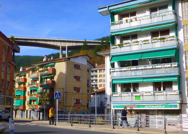 Eine Stadtstraßenszene mit Gebäuden, Fahrzeugen, Fußgängern, Straßenmöblierung, einer Brücke, fernen Hügeln und einem bewölkten Himmel.