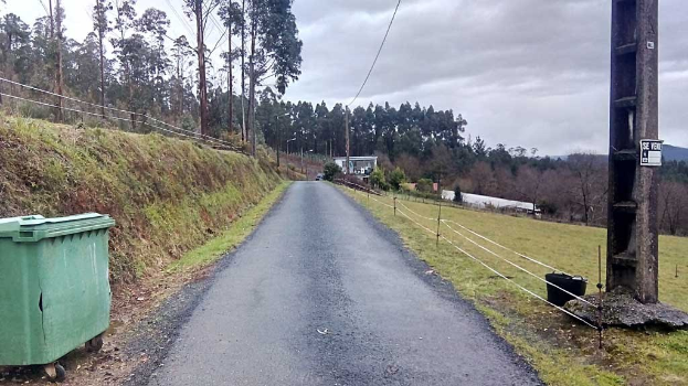 Grüner Müllcontainer auf der Seite einer Straße mit Gras, Pflanzen, Bäumen, Masten, Dröhnen und einem Zaun, unter einem bewölkten Himmel, mit einer Masttafel auf der rechten Seite und verstreuten Gegenständen auf der linken Seite.